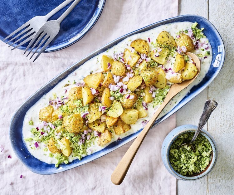 Een langwerpig blauw bord staat op tafel. Hierop is yoghurt op geschept met krokante aardappelen, stukjes rode ui en geraspte broccoli. 