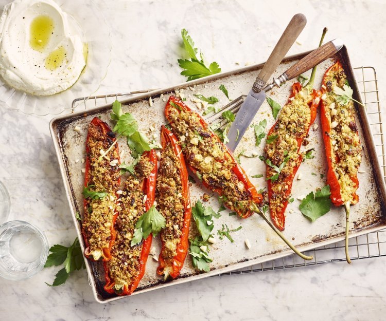 Gevulde puntpaprika's liggen op een metalen ovenschaal te schitteren. Ze zijn middendoorgesneden en opgevuld met couscous, gehakt en smaakmakers. Er liggen enkele blaadjes koriander bij voor extra kleur en het potje met homemade labneh is gepimpt met olijfolie en een draai van de pepermolen.