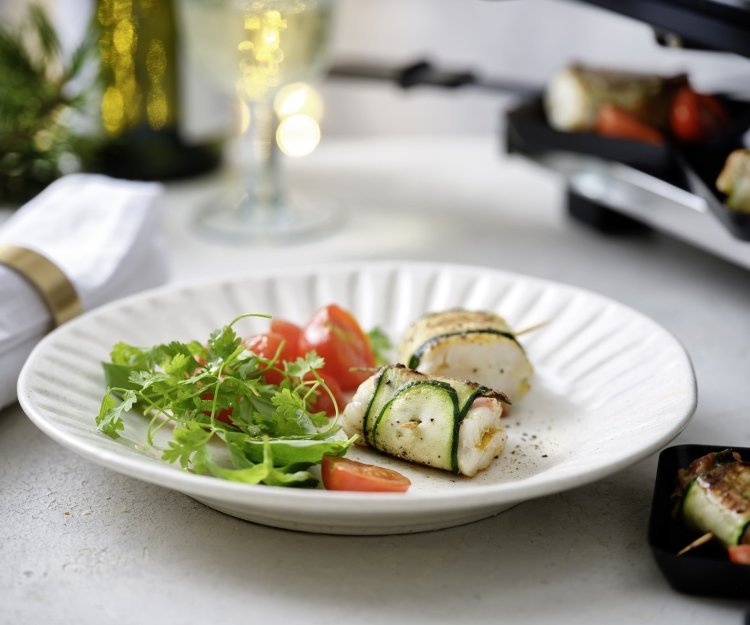 Twee pakjes liggen op een wit bord. Het pakje is gemaakt van een stukje vis verpakt in een sliertje courgette. We serveren dit feestelijk gerechtje met een frisgroene kruidensla en kertomaatjes.  