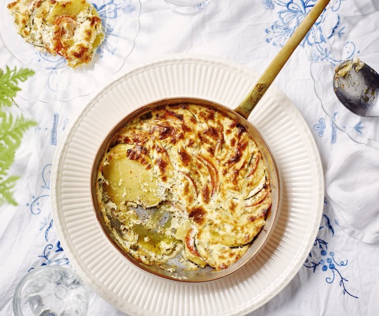 Een heerlijke gratin met groenten en kaas klaargemaakt in een ovenbestendige pan staat in het midden van de tafel. Er is al een portie uitgeschept op een bordje. Op tafel ligt een wit tafelkleed met enkele geborduurde blauwe bloemen. 