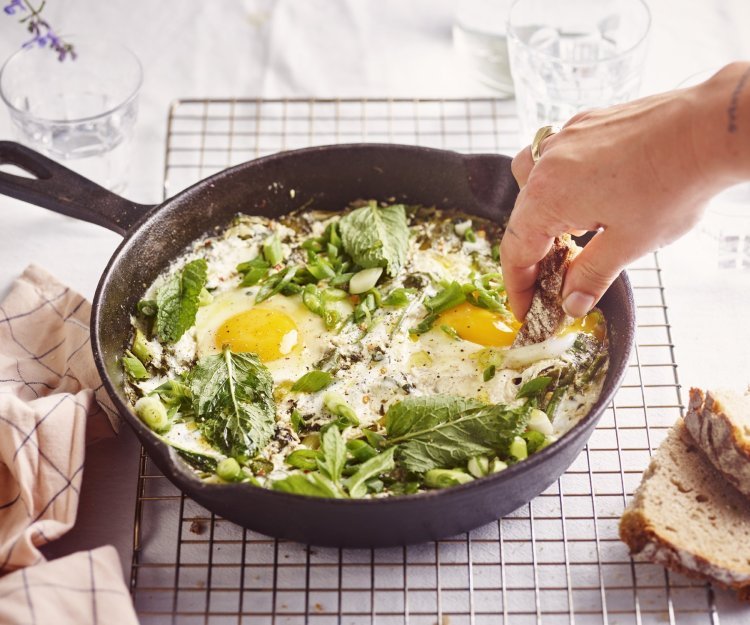 Op een rooster staat een zwarte gietijzeren pan met grieks getint gerechtje. Groene shakshuka met gebakken yoghurt. Er is een hand met een stukje brood in beeld dat er een dipje van zal nemen. 