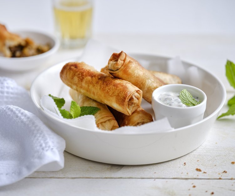 Meerdere rolletjes liggen te pronken in een wit serveerschaaltje. De rolletjes zien er heerlijk krokante gebakken uit en worden geserveerd met een dip van yoghurt en munt in een ander wit klein potje. Er ligt ook nog een takje munt bij voor een vleugje extra groen.