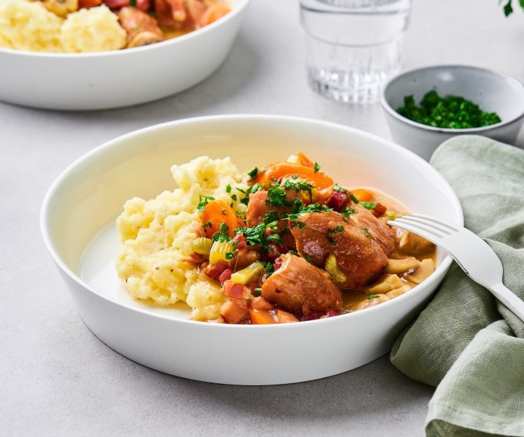 Op tafel staan twee uitgeschepte porties van het hoofdgerecht. Een schep heerlijke puree met kalfsfricassee. In de fricassee zitten spekblokjes, selder en wortelstukjes. Het gerechtje is afgewerkt met verse peterselie. Er ligt een licht groene servette langs en wit bestek.