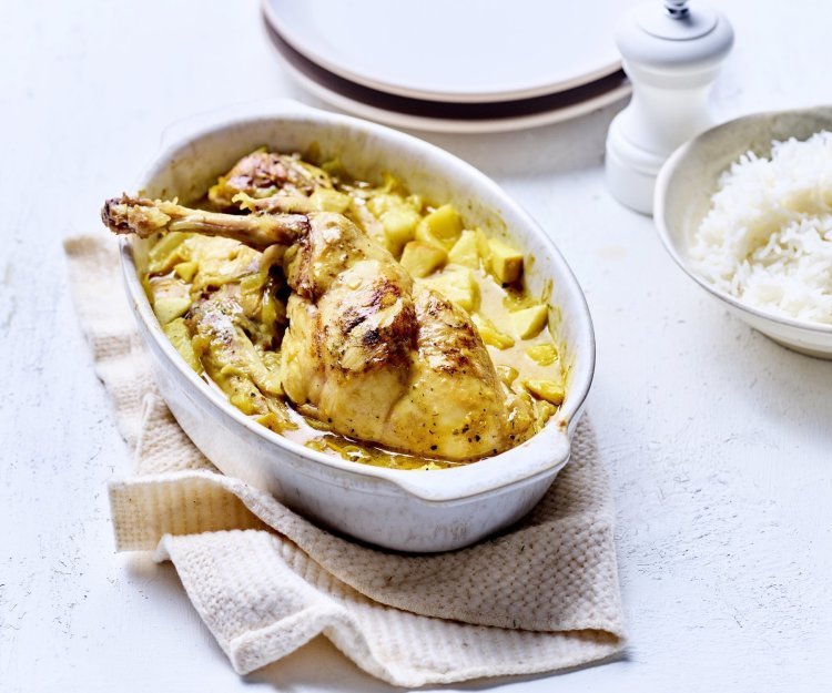 Op tafel staat een witte ovale ovenschotel met hierin de gele currysaus met stukjes appel en ananas. Hier bovenop liggen de bouten te blinken. De ovenschotel staat op een beige linnen handdoek met rijst, borden en een witte pepermolen langs.