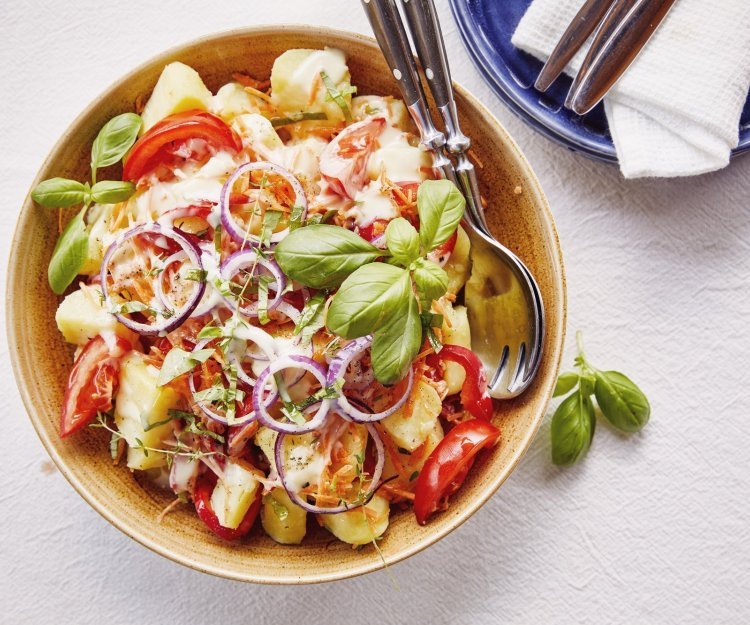 Een grote serveerschaal staat op de tafel, ze is gevuld met gekookte aardappelstukjes, tomaat, paparika, rode ui en wortel. De dressing is al toegevoegd en verse kruiden zoals basilicum en tijm geven een nog extra kleurtje aan de salade.