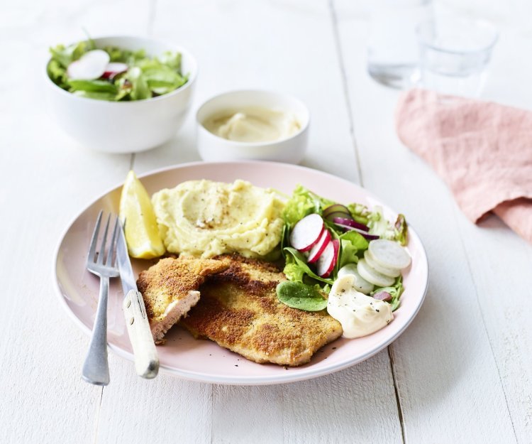 Een homemade shnitzel ligt op een pastelroze bord, samen met aardappelpuree, verse sla en zelfgemaakte mayonaise. Er is nog wat extra salade in een wit potje en mayonaise voor extra lekkers op te scheppen. 