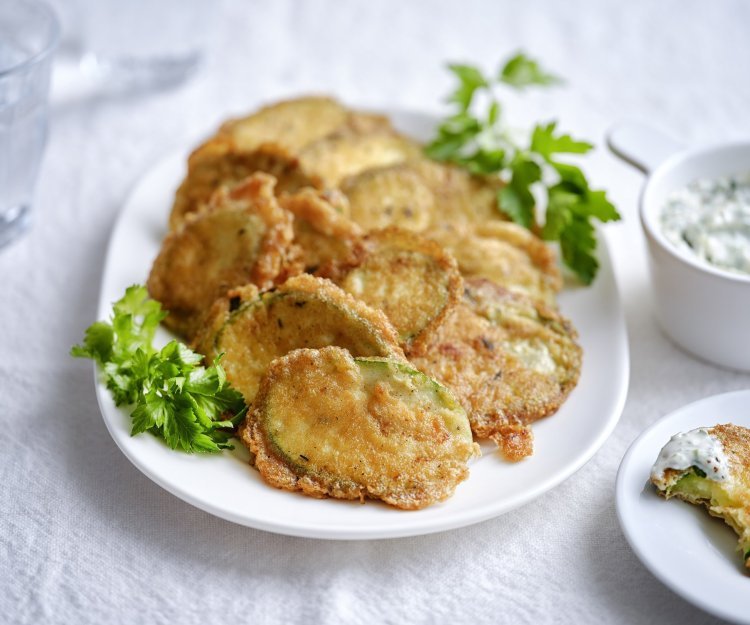 Krokante courgetteplakjes liggen op een bordje, afgewerkt met enkele blaadjes peterselie. De aioli staat langs het bordje.