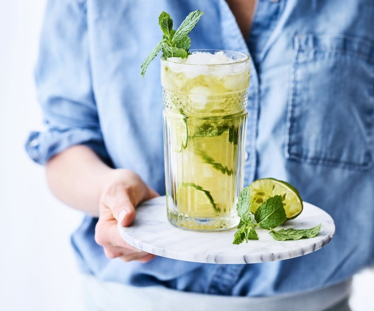 Een heerlijk frisse mocktail van Jupiler 0.0 en peach ice tea. Geserveerd met frisse limoenschijfjes in het glas. De vrouw draagt een blauw jeanshemd en staat mee in beeld.