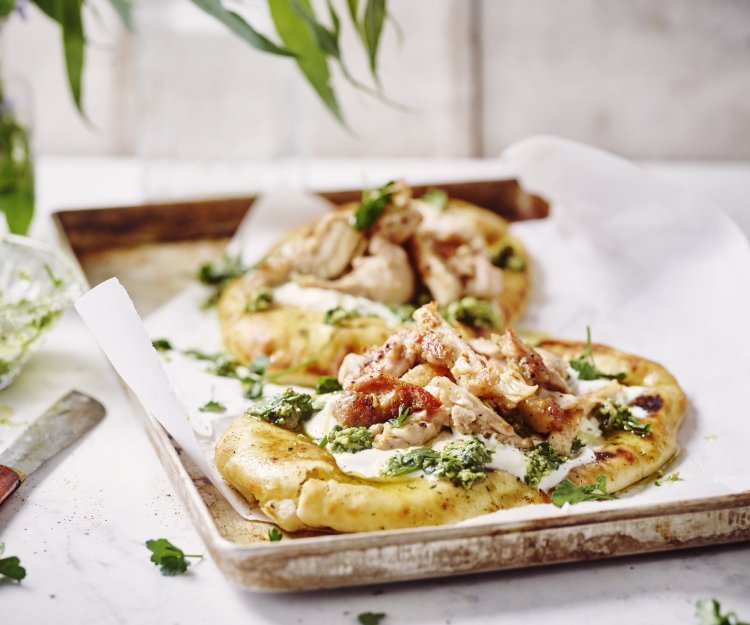 Twee naanbroodjes liggen op een metalen ovenplaat. Ze zijn belegd met yoghurt, stukjes gebakken kip en afgewerkt met salsa verde. De ovenplaat staat op een witte houten tafel.