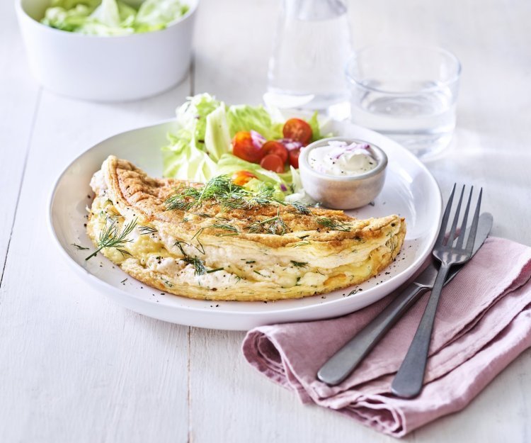 Een heerlijke omelet ligt op een wit bord. Het is een luchtige omelette die we combineren met verse kruiden en kaas. We voegde ook nog wat sla en tomaatjes toe aan het bord voor een extra portie kleur en groenten. 