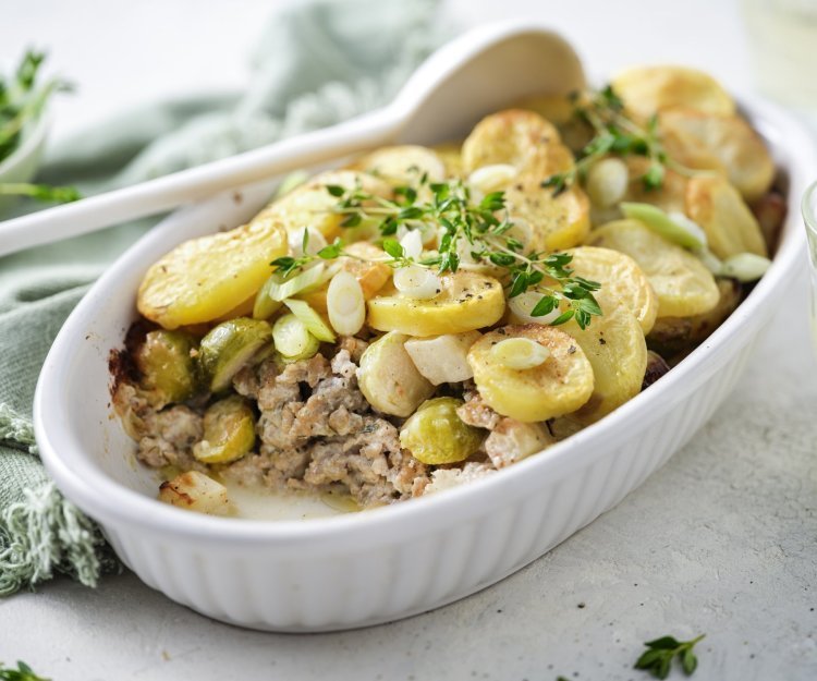 Een ovenschotel staat op het midden van de tafel, er is al een portie uitgeschept. Je ziet het gehakt onderaan liggen, met spruitjes , stukjes knolselder en goudgele aardappelschijfjes. 