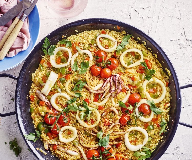 Een grote pan gevuld met een kleurrijke paella staat op het aanrecht. Hij zit vol met de goudgele rijst, in ringen gesneden pijlinktvis, kerstomaten, paprika en verse peterselie.  