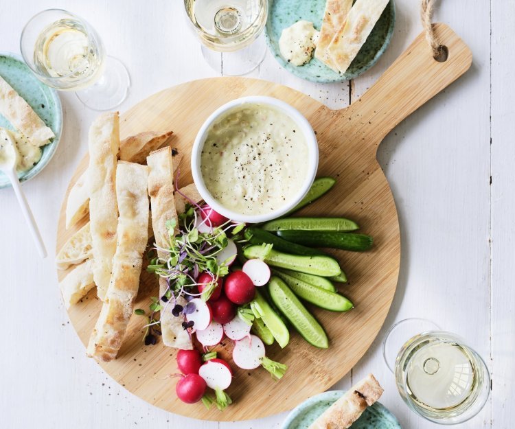 Op tafel staat een ronde houten snijplank met hierop een wit potje gevuld met onze pikante dip van yoghurt en witte bonen. Je kan de yoghurtdip gebruiken met stukjes snackkomkommer, brood en radijsjes in ons geval. Rond de plank staan enkele glazen witte wijn en helderblauwe broodbordjes. 