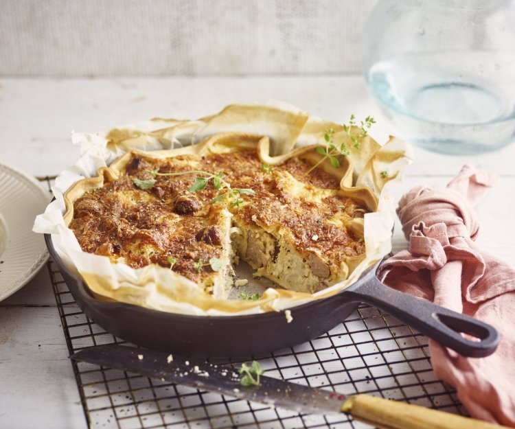 Op het aanrecht ligt een zwarte rooster met hierop de quiche in een gietijzeren pan. Hierbij ligt een roze servet, mes en bordje waarop al een stukje quiche op ligt. De quiche is afgewerkt met enkele verse blaadjes oregano. 