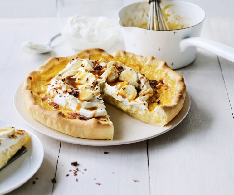 Op een witte houten tafel staat een klaargemaakte rijsttaart, hij is afgewerkt met vers opgeklopte slagroom, stukjes banaan en vloeibare karamel. Er is al een stukje aangesneden. Verder uit beeld staat het steelpannetje waarin de karamelsaus is klaargemaakt.