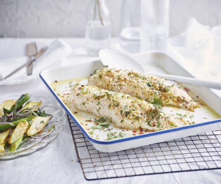 De gegrilde roodbaars uit de oven ligt op een witte bakplaat en staat op een rooster. De crumble zorgt voor een zomerse sfeer in het gerecht. Naast de ovenschaal staat een bordje met wat aardappelsalade uitgeschept. 