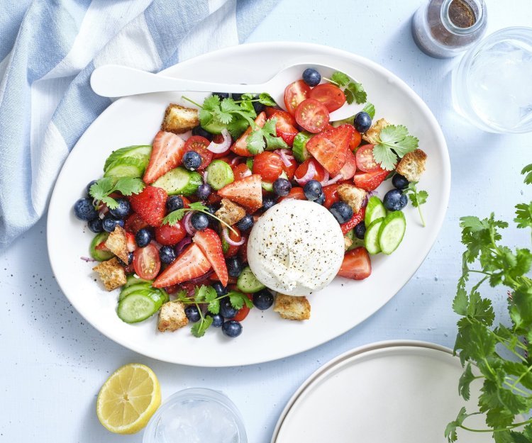 Er staat een grote schaal op tafel boordevol zomerse toppers. Denk maar aan tomaatjes, aardbeien, bosbessen, komkommers en burrata. Dit geeft echt verfrissing tijdens een zwoele zomerdag. Er ligt een citroentje langs, een glas water en verse kroiander.