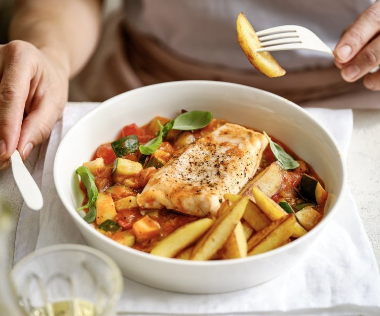 Aan taffel zit een vrouw die een stukje aardappel prikte op haar vork. Voor haar staat een bordje gevuld met het gerecht, vis met Prvençaalse saus en gebakken aardappelpartjes. 