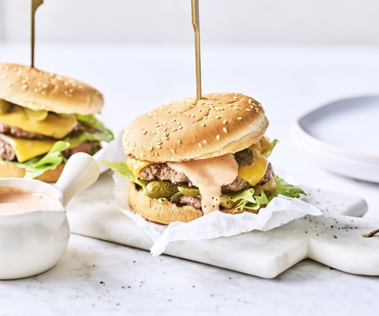 Twee heerlijke burgers liggen klaar op een marmere snijplank. Het vlees is verdeeld in twee dunne porties met de saus en gesmolten kaas er bovenop. De burgers worden recht gehouden door middel van een prikker.