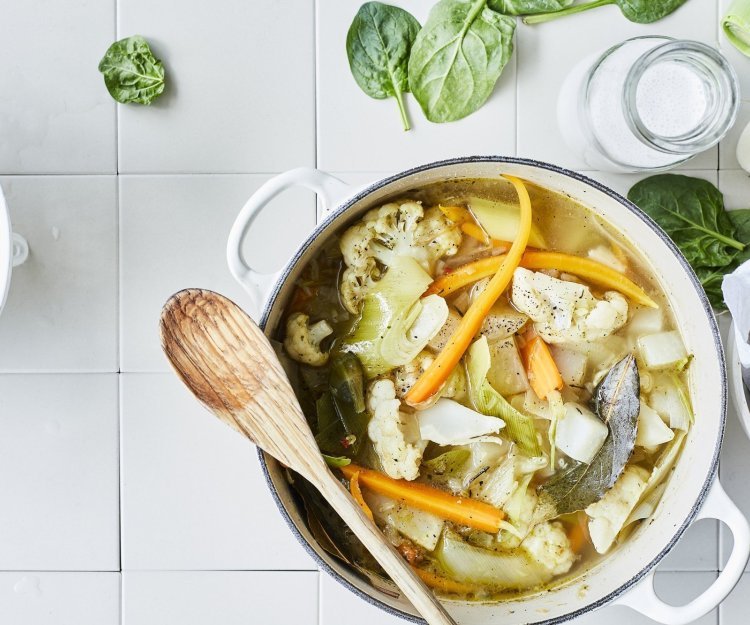 Een lentestoofpotje boordevol groenten, denk maar aan bloemkool, wortel, koolrabi, prei en spinazie. De stoofpot staat op het aanrecht in een grote kookpot, rond de pot liggen nog enkele spinazieblaadjes en een flesje room.