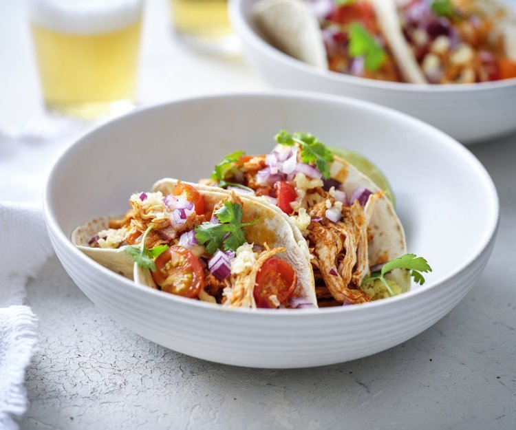 In een diep wit bord liggen twee gevulde taco's met pulled chicken, afgewerkt met tomaat, rode ui en geraspte Passendael kaas. Om wat extra kleur te geven liggen er enkele blaadjes koriander op. Wij serveren ze met frisgekoeld biertje, smakelijk!