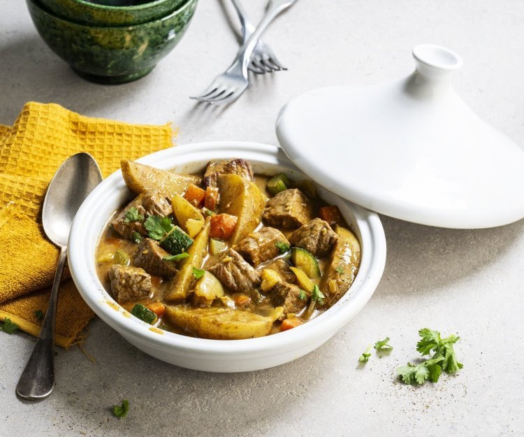 Op tafel staat een witte tajine met een gerechtje van rundvlees, courgette, selder, wortel en stukjes aardappel. Er ligt een okerkleurige servette bij en groene kommetjes om de tajine in op te scheppen. 