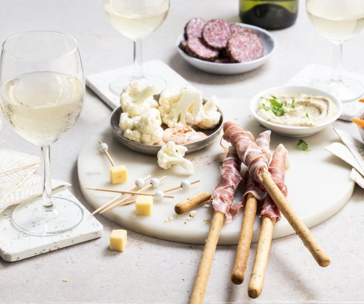 Een mooie marmere serveerplank in rond vorm staat op tafel. Hij is gevuld met allerlei tappas, zoals bloemkoolroosjes met cocktailsaus, wortel, kaasblokjes, salami met mosterd en homemade hummus.