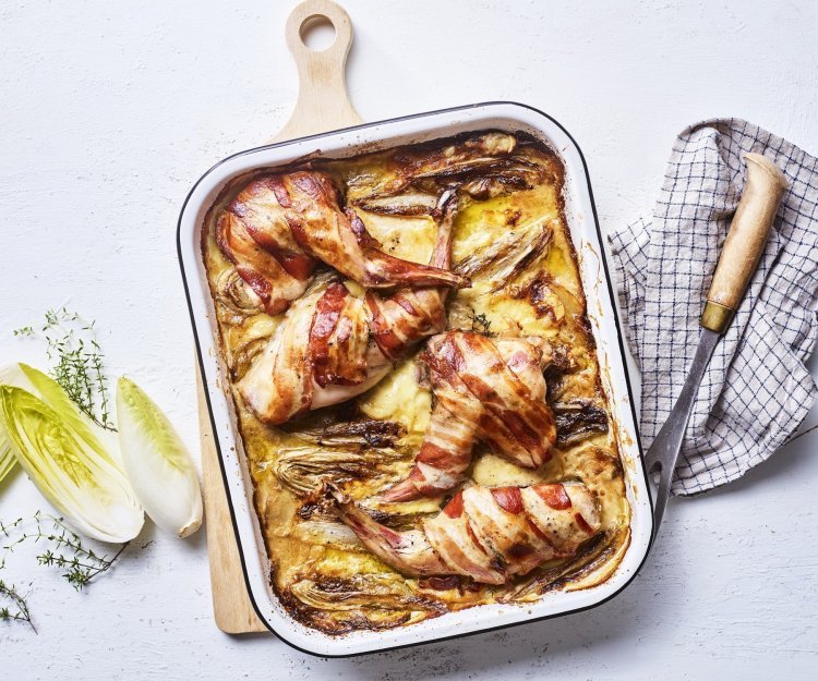 Op een grote houten snijplank staat de ovenschaal met de tartiflette en stukken konijn. Het konijn in omwikkeld met sneetjes spek, waardoor het nog iets extra heeft. Er ligt witloof langs en verse tijm.