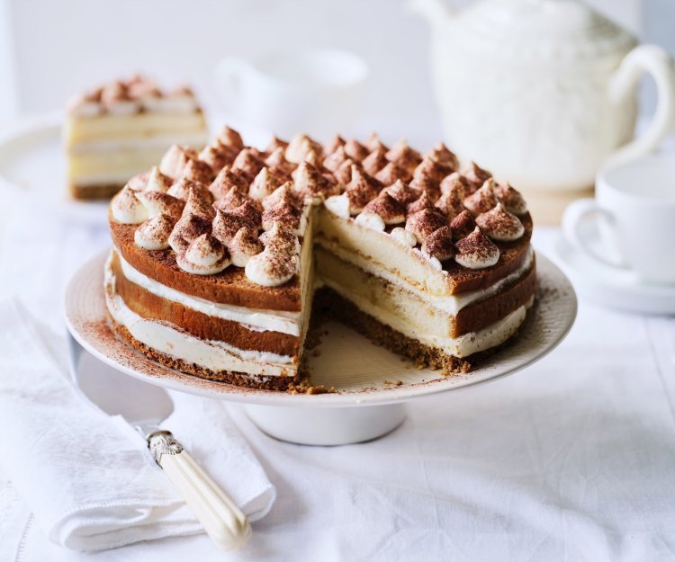 Op tafel staat een prachtige taart, het is een twist op de klassieke tiramisu. Hij staat op een witte taartschaal, en is afgewerkt met toefjes mascarpone en cacaopoeder. In de achtergrond staat een koffiekan en tasjes koffie.