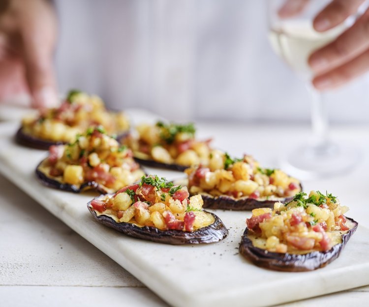 Op een marmere serveerschaal liggen enkele aubergineplakjes. Deze dienen als toastje en zijn belegd met stukjes Berloumi en spek. 