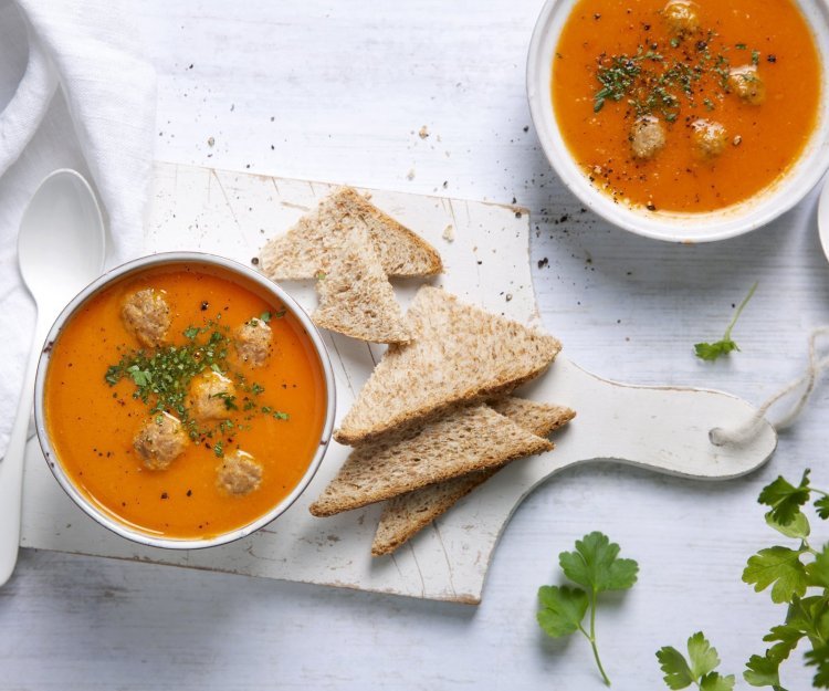 Heerlijk gemaakte tomatensoep staat uitgeschenkt op tafel in twee soepborden. De balletjes drijven in de soep en is afgewerkt met peterselie. Brood en lepels liggen langs het bord, klaar om te slurpen.