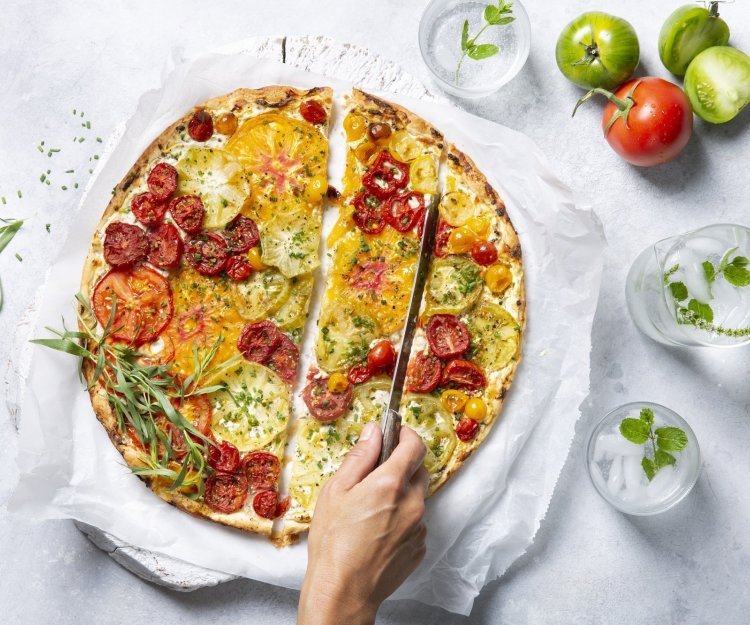 Een kleurrijke tomatentaart staat op tafel op een wit boterpapiertje. Ze wordt aangesneden door een onbekende vrouw met een koksmes. Rechtsboven liggen nog enkele tomaten om ze extra in de picture te zetten. 