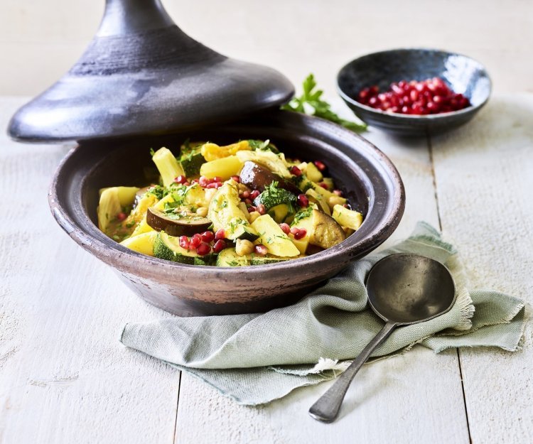 Er staat een tajine op tafel, de tajine heeft een zwarte kleur en is gevuld met stukjes aardappel, courgette, venkel, abrikozen, kikkererwten en granaatappelpitten. 