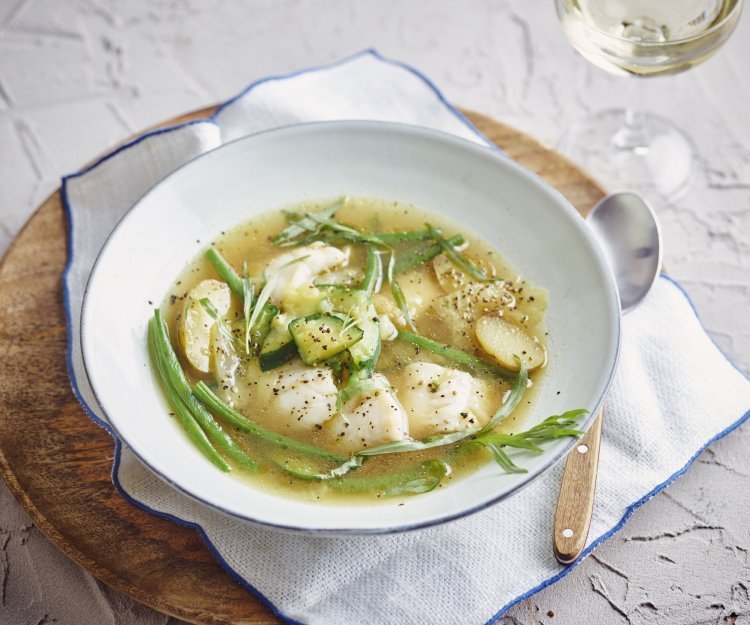 Een heerlijke maaltijdsoep is geserveerd in een wit soepbord. Deze staat op tafel, op een houte ronde snijplank, op een witte servette met blauwe rand. Het bord is gevuld met de doorschijnende bouillon met hierin de stukjes groenten en vis. Echt een feestsoepje. 