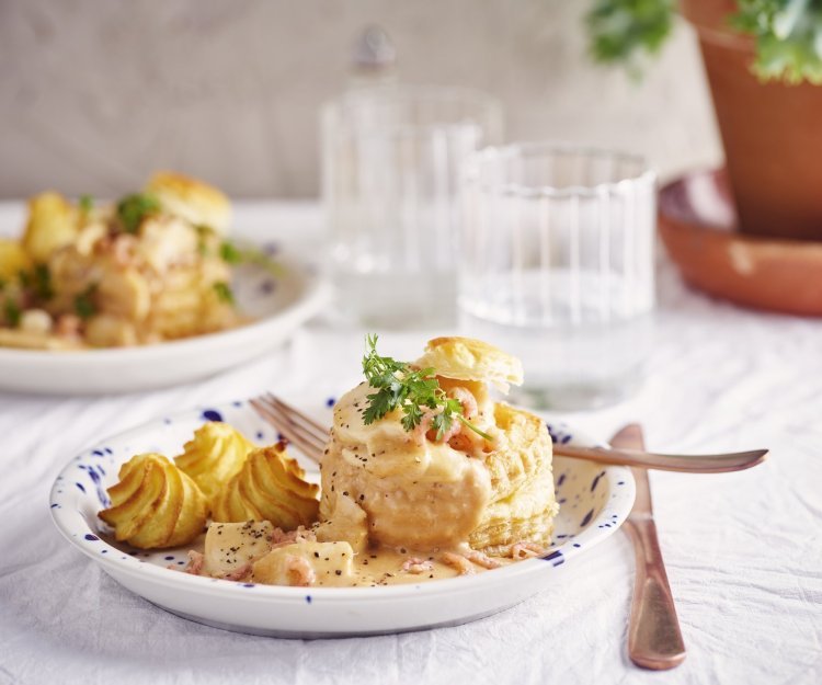 Twee borden staan klaar om van te smullen. Vol-au-vent van vis in een bladerdeegpasteitje met pommes duchesse. Er staat een glaasje water naast het bord.