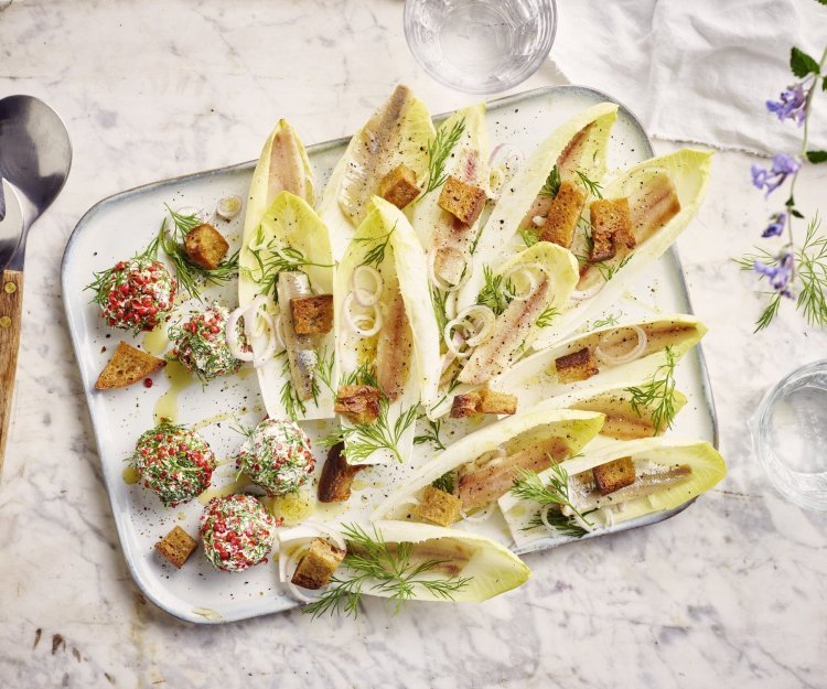 Op een rechthoekige dienschotel liggen meerdere blaadjes van witloof gevuld met een stukje sprot, ringen van sjalot en croutons. In het hoekje van de schotel liggen de roomkaasballetjes, deze balletjes zijn gerold in roze peperkorrels en verse dille waardoor ze als het ware een jasje van kruiden kregen.