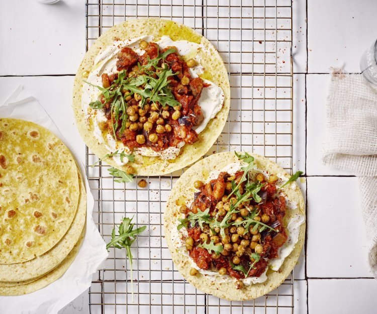 Twee wraps liggen op een ovenrooster klaargemaakt. Ze zijn belegd met roomkaas, tomatenspread en geroosterde kikkererwten. Er liggen nog enkele extra wraps langs. Het gerechtje is afgewerkt met enkele blaadjes rucola.