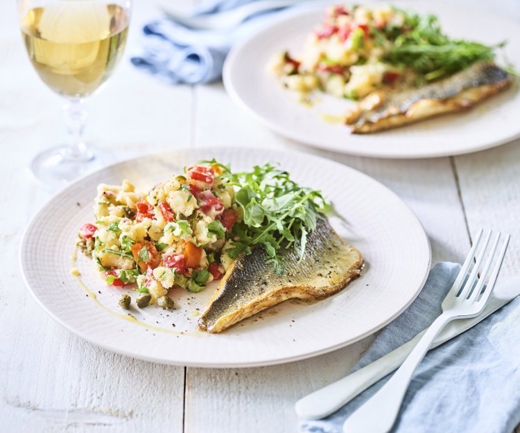 Op tafel staan twee borden met het gerechtje op. Het is een aardappelpuree met stukjes paprika, kappertjes en rucolasla. En een krokant gebakken zeebaarsfilet. Er staat ook een glaasje witte wijn langs. 