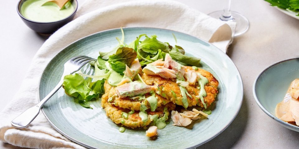 Op tafel staat een licht blauw bord, met hierop de goudgele pannekoekjes op basis van aardappelpuree. Het gerechtje is afgewerkt met enkele stukjes gerookte forel, sla en een groene dressing van verse kruiden. 