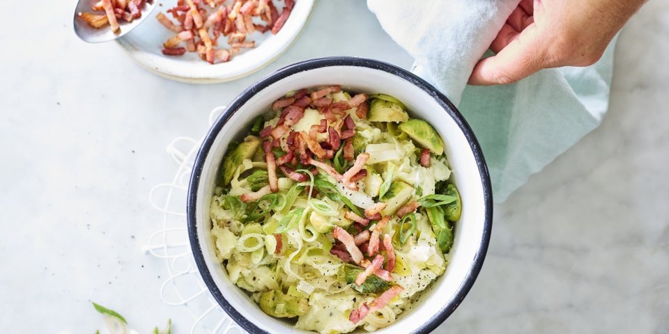 Smeuïge aardappelpuree met grofgehakte spruitjes en prei klaargemaakt in een kookpot. De kok in kwestie voegt de versneden lente-uitjes en gebakken spekjes toe als afwerking.