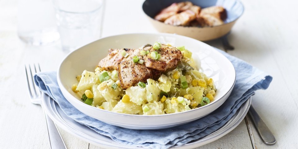 Een rijkgevulde aardappelsalade is opgediend in een diep wit bord. Hier bovenop ligt het krokant gebakken spek. De overige stukjes spek liggen nog in het bakpannetje en staat bovenaan het bord in beeld. 