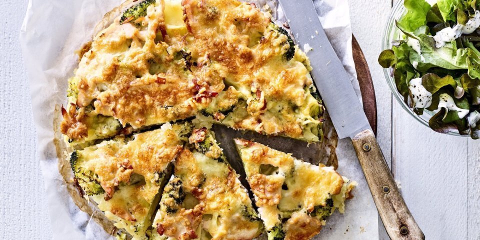 Deze hartige taart lijkt op een quiche maar is toch anders. Deze is gemaakt van aardappelschijfjes, broccoli en kippenwit. Ze ligt op een houten ronde snijplank met boterpapiertje onder, ze is al voor de helft aangesneden. Rechtsboven staat nog een kommetje gemengde sla. 