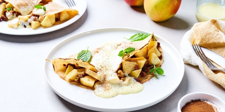 Op tafel staan twee borden met elk hierop een dichtgevouwen pannenkoek, die gevuld is met stukjes appel en rozijnen. Wij serveren hem met vanillesaus en verse blaadjes munt. Er liggen ook nog enkele appelen in de achtergrond en een restje vanillesaus.