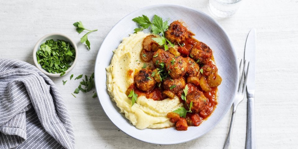 Een licht grijs bord staat op een witte tafel. Hierop is een schep smeuïge puree open geschept met daarbij de balletjes in tomatensaus. Het gerechtje is afgewerkt met wat verse peterselie. 