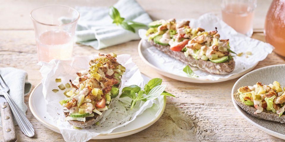 Er staan 3 bordjes klaar op een houten tafel. De ideale lunch om te benuttigen buiten in de zon. De halve broodjes zijn belegd met kruidige kip en verse groentjes. 