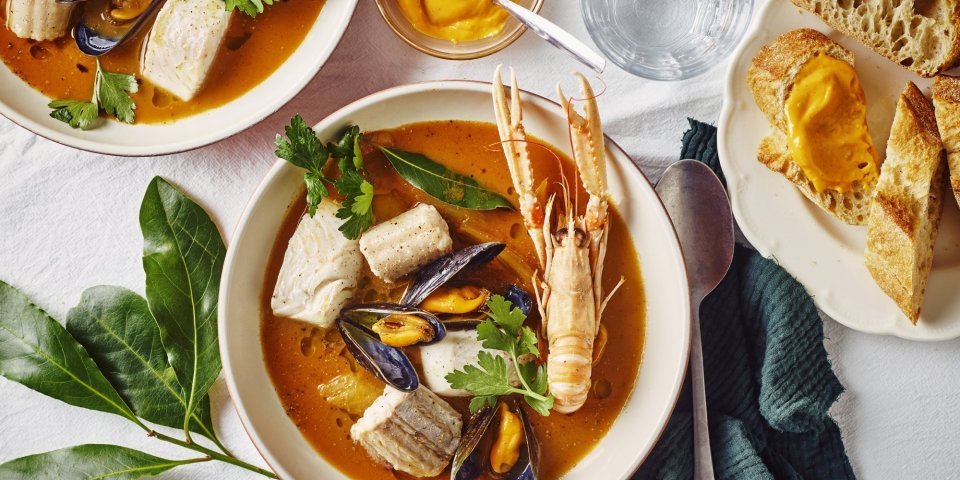 Een heerlijk kleurrijk bordje bouillabaisse staat op tafel. Wij maakten hem met topproducten van hier. Er ligt een donker groene linnen servette langs en bordje stukjes brood en verse rouille.