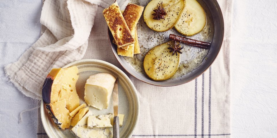 Op tafel ligt een handdoek opengevouwen met hierop een bord en een pan. Op het bord ligt een verzameling Belgische kazen en een mes, in de pan liggen gekaramiliseerde peren, safraanstokje, steranijs en briochetoastjes. 