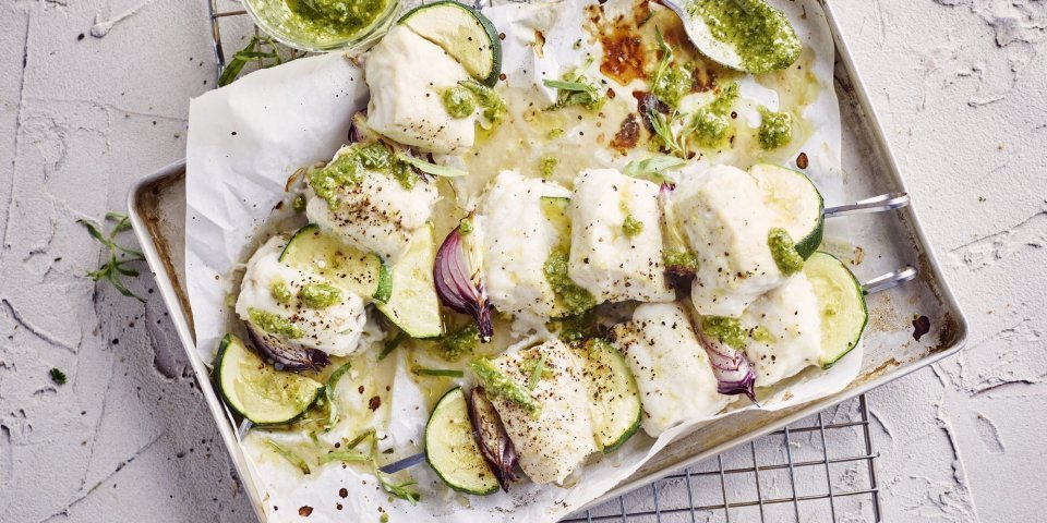 Klaargemaakte brochettes liggen in een grijze ovenschaal te blinken. De spiesjes bestaan uit stukjes heek, courgette en rode ui. Er staat een glazen potje naast met huisgemaakte dragonpesto.