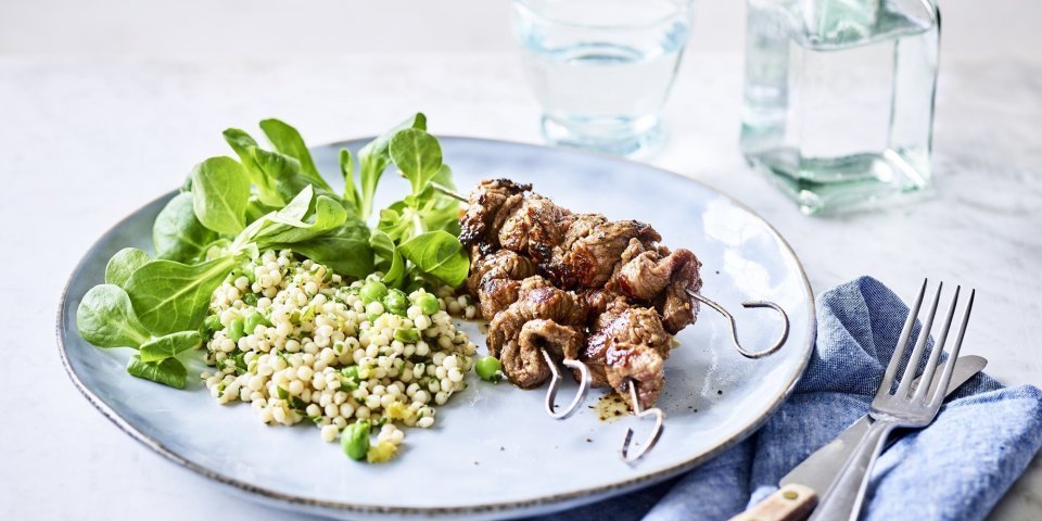 Op een lichtblauw groot bord liggen 3 gegrilde brochettes, we serveren ze met een couscoussalade van erwtjes en verse kruiden. Naast het bord ligt een blauwe servette met bestek langs. Er staat ook een glaasje water bovenaan het bord met een flesje water naast.  