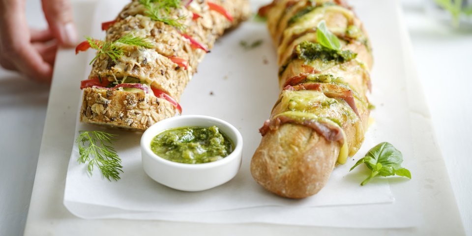 Twee broodjes liggen op een snijplank. Je ziet dat het twee versies zijn, nl een broodje met groene pesto, sneetjes ham en kaas. En dan nog een versie met roomkaas, paprika en rode ui. Extra pesto staat in een potje bij de broodjes, voor wie wat extra wil. Een vrouw zet de plateau of snijplank op tafel, haar handen zijn nog in beeld. 